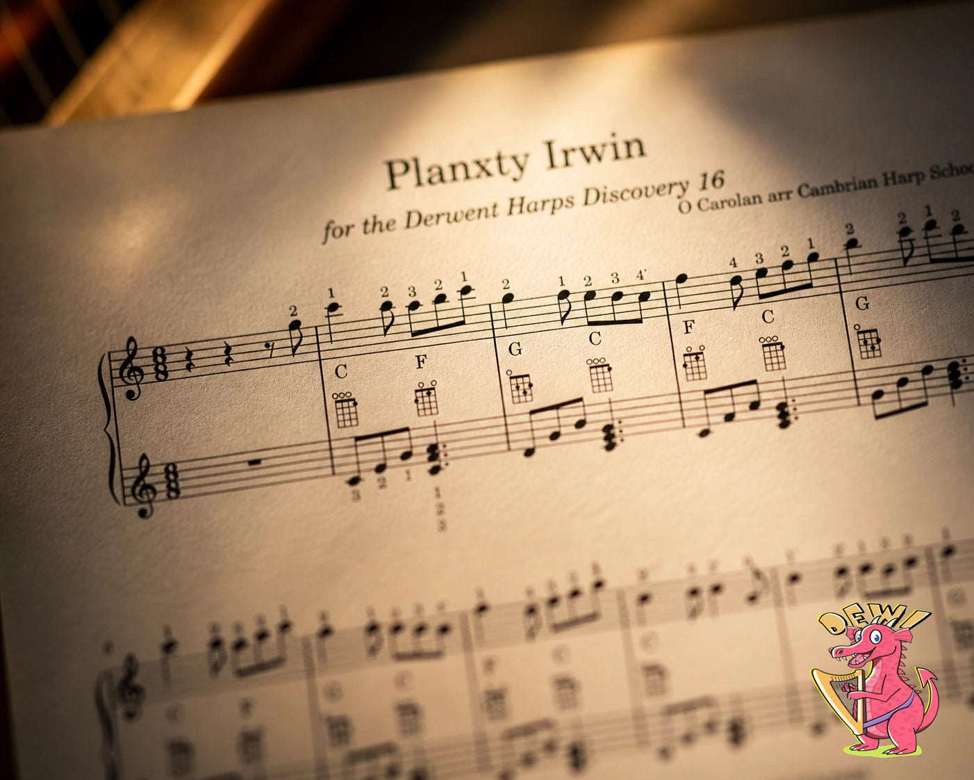 Close-up artistic photograph of sheet music for 'Planxty Irwin' arranged for the Derwent Harps Discovery 16, showing musical notation, fingerings, and ukulele chord diagrams under warm, focused lighting with a small pink dragon mascot in the corner.