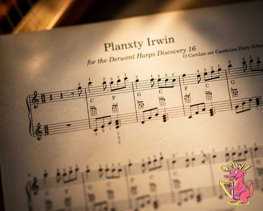 Close-up artistic photograph of sheet music for 'Planxty Irwin' arranged for the Derwent Harps Discovery 16, showing musical notation, fingerings, and ukulele chord diagrams under warm, focused lighting with a small pink dragon mascot in the corner.
